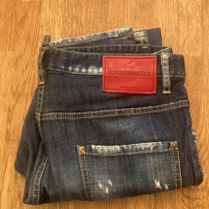Mörkblå Dsquared2 jeans med slitningar - Snygga mörkblå jeans från Dsquared2 med coola slitningar och detaljerad tvätt. Klassisk femficksmodell med kontrastsömmar och röd Dsquared2-logga på bakfickan och vid gylfen. Jeansen har rak passform och är tillverkade i robust denim.