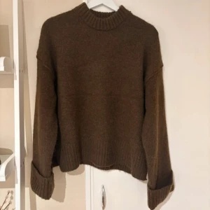 Brun stickad tröja från Gina Tricot - Mysig brun stickad tröja från Gina Tricot med ribbad hals och breda muddar vid ärmsluten. Tröjan har en loose fit och är perfekt för lager-på-lager under kyliga dagar. Enkel att matcha med jeans eller kjol för en chill look.