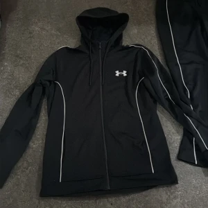 Svart träningsset från Under Armour - Säljer ett svart träningsset från Under Armour med vit logga och tunna vita linjer längs ärmar och ben. Hoodien har dragkedja och huva, byxorna har resår i midjan och smal passform. Perfekt för dig som gillar sportig och stilren look.