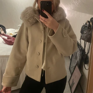 Beige kappa med fluffig krage - Snygg beige kappa med stor fluffig krage i pälsimitation och klassiska knappar framtill. Jackan har en stilren och rak passform, perfekt för dig som gillar en clean look. Pälsen på luvan är avtagbar