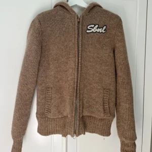 Brun stickad jacka från Sissy-Boy - Mysig brun stickad jacka från Sissy-Boy med dragkedja och broderad text på bröstet. Jackan har huva fodrad med fluffigt fuskpälsmaterial och två fickor framtill. Den är i storlek M men skulle säga att den mer passar storlek S. Perfekt för kyliga dagar och chill vibes.
