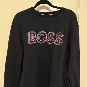 Svart sweatshirt från BOSS med tryck - Svart sweatshirt från BOSS med stort, broderat BOSS-tryck i vitt, rött och grönt på bröstet. Rund hals med ribbad kant och färgade ränder i grått, rött och mint. Långärmad och tillverkad i mjuk bomull för skön komfort.