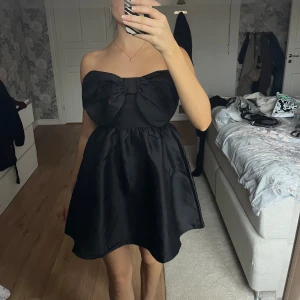 Svart axelbandslös klänning med rosett - Supersöt svart axelbandslös klänning med stor rosett framtill. Kjolen är kort och har en lätt utsvängd form som ger en fin siluett. Materialet är glansigt och känns lyxigt, perfekt för att sticka ut. Klänningen är helt ärmlös och har en stilren look.