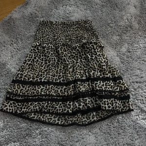 Leopardmönstrad kjol från Lindex - Snygg leopardmönstrad kjol från Lindex med smock i midjan och volanger. Kjolen har svarta spetsdetaljer och är i en luftig, lätt kvalitet. Perfekt för dig som vill sticka ut med en trendig look.