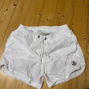 Moncler badbyxor  - Snygga vita badshorts från Moncler med elastisk midja och snörning framtill. Shortsen har en bakficka med dragkedja och Moncler-logga på vänster ben. Perfekta för sommaren och strandhäng!