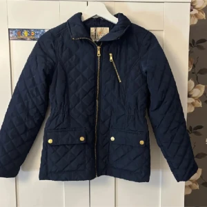 Mörkblå quiltad jacka Holly & White - Quiltad mörkblå jacka från Holly & White med guldfärgade dragkedjor och knappar. Jackan har två stora fickor framtill, en sned bröstficka med dragkedja och markerad midja. Perfekt för dig som gillar stilen och klassisk look. Sår Stella på insidan av jackan. Skriv för fler bilder!