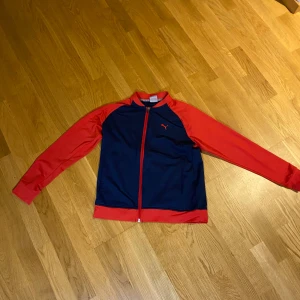 Blå och röd träningsjacka från Puma - Sportig träningsjacka från Puma i marinblått med röda ärmar och detaljer. Jackan har hel dragkedja framtill, ribbad krage och muddar samt Puma-logga på bröstet. Tillverkad i mjuk polyester som passar perfekt för träning eller chill.