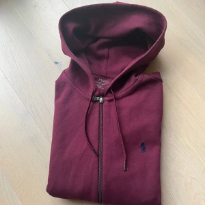 Vinröd hoodie från Polo Ralph Lauren - Snygg vinröd hoodie från Polo Ralph Lauren med dragkedja och huva. Tröjan har långa ärmar, två fickor fram och den klassiska Polo-loggan broderad på bröstet. Perfekt för chill dagar och enkel att matcha med jeans eller joggers.