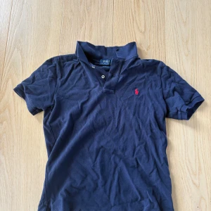 Marinblå pikétröja från Ralph Lauren - Klassisk marinblå pikétröja från Ralph Lauren med röd broderad logga på bröstet. Tröjan har korta ärmar, krage och två knappar framtill. Tillverkad i bomull för en skön och avslappnad känsla.