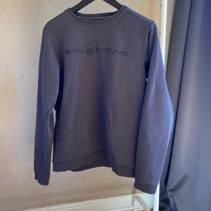 Mörkgrå Sail Racing sweatshirt - Säljer en mörkgrå sweatshirt från Sail Racing med tryckt logga på bröstet. Tröjan har rund halsringning, långa ärmar och är tillverkad i mjuk bomullsmix. Perfekt för chill dagar eller när du vill ha en clean look.