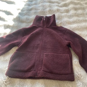 Vinröd teddyjacka med dragkedja - Mysig vinröd teddyjacka med hög krage och hel dragkedja framtill. Jackan har två stora fickor framtill och är tillverkad i ett fluffigt teddy-material som håller dig varm. Passformen är lite loose och ärmarna är långa, perfekt för kyliga dagar.