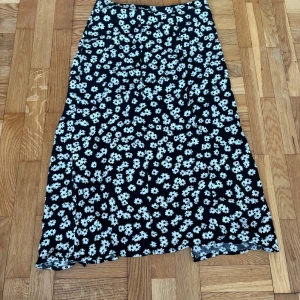 Svart blommig långkjol från Amisu - Svart långkjol från Amisu med vita blommor över hela tyget. Kjolen har en lätt utsvängd form och är perfekt för dig som gillar en feminin och trendig stil. Mjukt och följsamt material som känns skönt mot huden.