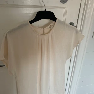 Ljusbeige blus från Vero Moda - Säljer en ljusbeige blus från Vero Moda med rund halsringning och korta ärmar. Blusen har ett lätt, skirt material och fina rynkdetaljer vid halsen. Perfekt för dig som gillar en enkel och stilren look.