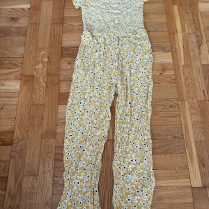 Gul blommig byxdress från Lindex - Superfin gul byxdress från Lindex med blommigt mönster i vitt och svart. Byxdressen har smock upptill, korta ärmar och ett flowigt fall. Perfekt för dig som gillar färg och mönster! 