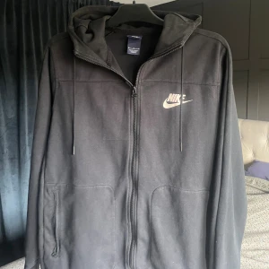 Svart Nike hoodie med dragkedja - Svart hoodie från Nike med vit logga på bröstet. Tröjan har huva med snörning, hel dragkedja framtill och två fickor med dragkedja. Tillverkad i mjuk bomullsblandning och har lång ärm. Perfekt för en chill och avslappnad look.
