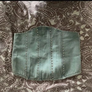 Ljusblå denim korsett från Boohoo - Säljer en ljusblå denim korsett från Boohoo med silverfärgade nitar i vertikala rader framtill. Toppen har en snygg formad överkant och dragkedja bak för enkel påklädning. Perfekt för att ge din outfit en edgy vibe.