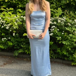 Ljusblå långklänning med öppen rygg - Supersnygg ljusblå långklänning i satinliknande material med tunna axelband och öppen rygg. Klänningen har en figurnära passform, drapering på sidan och dragkedja bak. 🩵