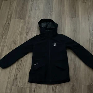 Svart vindjacka från Haglöfs Junior - Svart vindjacka från Haglöfs med huva och dragkedja framtill. Jackan har två snedställda fickor med lock och diskret Haglöfs-logga på bröstet. Tillverkad i slitstarkt syntetmaterial och har meshfoder på insidan. Perfekt för blåsiga dagar.