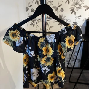 Offshoulder topp! - Superfin offshoulder topp från Gina Tricot i svart med stora gula och vita blommor. Volang upptill och elastisk kant som håller toppen på plats. Perfekt för sommarens alla tillfällen och lätt att matcha med jeans eller kjol. Såå fin men tycker den är för kort på mig!