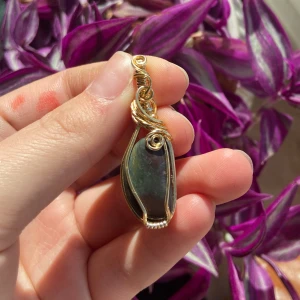 Guldberlock med grön och lila sten   - Unik berlock med en mörkgrön, slät sten infattad i snirklig guldtråd. Plockad på Sicilien och polerad av mig.  Hängsmycket har en organisk form och detaljerade trådvirvlar som ger en cool och bohemisk vibe. Perfekt för dig som gillar naturinspirerade smycken och vill sticka ut med något personligt. Kedja kostar 50kr extra. 