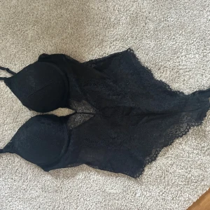 Svart spetsig body från hm!  - Säljer min svarta spets body från hm i genombruten spets med vadderade kupor och tunna axelband. Toppen har en feminin och elegant look med fina spetsdetaljer och transparenta partier. Perfekt för att lyfta din outfit med en edgy touch. Strl 80 C
