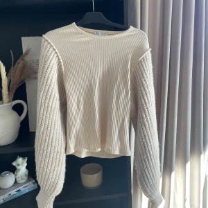 Beige ribbad tröja från Zara - Säljer en beige ribbad tröja från Zara med stickade, fluffiga ärmar och rund halsringning. Tröjan har en snygg kontrast mellan den ribbade kroppen och de grovstickade ärmarna. Storlek M