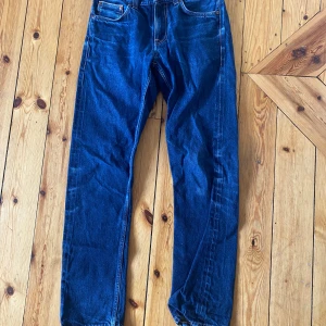 Nudie jeans grim trim - Säljer ett par klassiska blå jeans från Nudie Jeans Co med snygga kontrastsömmar och ikonisk broderad bakficka. Jeansen har normal passform, raka ben och är tillverkade i slitstarkt bomullsjeans. Perfekta för dig som gillar tidlös stil och bra kvalitet.