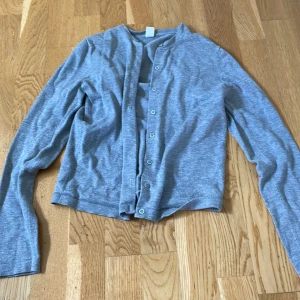 Grå kofta från Å Woman XS - Ljusgrå kofta från Å Woman i storlek XS. Koftan har långa ärmar, rund halsringning och knäppning framtill med små knappar. Tillverkad i mjukt bomullsmaterial, perfekt för lager-på-lager och chill dagar.