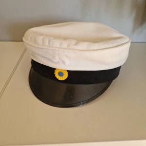 Studentmössa  - Klassisk studentmössa med vit ovandel i bomull, svart band och blank svart skärm (troligtvis skinn).  Insidan har foder i blått och gult tyg samt brunt svettband. Storlek 54, vilket motsvarar ca. 115 från ABC-gruppen.