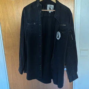 Svart overshirt - Svart overshirt från Alan Rust med dragkedjedetalj på ena ärmen. Skjortan har knappar framtill, bröstficka och en avslappnad passform. Aldrig använd