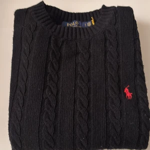 Svart kabelstickad tröja Polo Ralph Lauren - Kabelstickad svart tröja från Polo Ralph Lauren med klassisk röd broderad logga på bröstet. Tröjan har rund halsringning, ribbade muddar och är långärmad. Perfekt för dig som gillar stilren och tidlös design.
