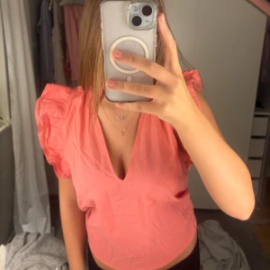 Korallrosa blus med volangärmar H&M - Superfin korallrosa blus från H&M med djup v-ringning och volangdetaljer på axlarna. Blusen är ärmlös och har en luftig passform, perfekt för varmare dagar. Tillverkad i mjuk bomull som känns skön mot huden.
