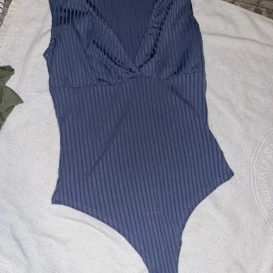 Bodysuit - Säljer en ribbad mörkblå bodysuit från NLY One i storlek XS. Bodyn har djup v-ringning och är ärmlös, perfekt att styla med jeans eller kjol. Materialet är stretchigt och följsamt för en tight passform.