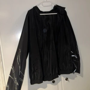 Vindjacka med reflex - Svart vindjacka från Zizzi med huva och dragkedja framtill. Jackan har coola reflexer detaljer längs ärmarna som ger en unik look. Perfekt för blåsiga dagar och för träning. Elastiska muddar vid ärmslut och nederkant för skön passform.