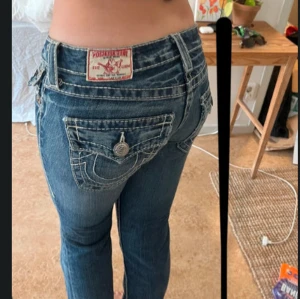 True Religion Joey Low Rise Flare Jeans - Säljer ett par blå True Religion Joey jeans med låg midja och flare-ben. Klassiska kontrastsömmar, stora fickor med lock och snygga detaljer i silver. Jeansen är i denim med tydliga gula och vita sömmar samt ikoniska knappar och patch bak. Avklippa förhand längst ner. Passar någon runt 160 