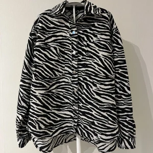 Oversized zebramönstrad skjortjacka - Säljer en tunn oversized skjortjacka med svartvitt zebramönster och stora bröstfickor. Jackan har en klassisk krage, knäppning framtill och långa ärmar. Materialet känns som tunnare jeanstyg. Varan har inga tecken på användning. 