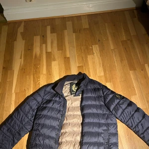 Blå pufferjacka från Belstaff - Snygg svart pufferjacka från Belstaff med quiltad design och guldfärgad logga på ärmen. Jackan har dragkedja framtill, ribbad krage och beige insida. Perfekt för dig som vill ha en stilren och varm jacka med streetkänsla.