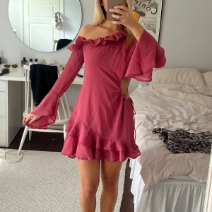 Rosa/röd offshoulder volangklänning - Superfin klänning med offshoulder och volangdetaljer. Klänningen har en snygg öppen detalj vid midjan. 