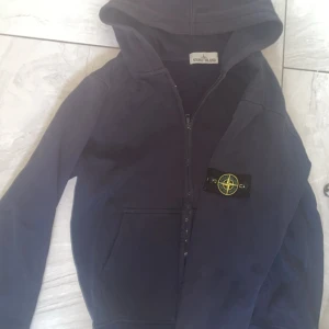 Mörkblå hoodie från Stone Island - Säljer en mörkblå hoodie från Stone Island med dragkedja och klassisk logga på ärmen. Tröjan har huva, känguruficka och är perfekt för dig som gillar streetwear. Grym passform och stilren design.