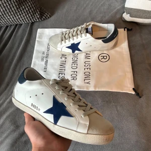 Golden goose 41 - Golden Goose Superstar sneakers i vitt skinn med blå stjärna på sidan och blå hälkappa. Grå mockadetaljer på tån och grå snörning. Klassisk låg modell med platt sula och rund tå. Snygg kontrast mellan färgerna och ikonisk branding på häl och plös. skriv vid frågor priset går att diskutera 