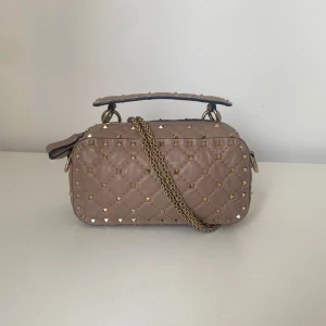 Valentino Garavani Rockstud Spike Free handväska - Snygg beige axelväska från Valentino med quiltat mönster och guldiga nitar. Väskan har kedjerem, dragkedja och ett stadigt handtag upptill. Tillverkad i mjukt skinn och har en lyxig vibe som verkligen sticker ut.