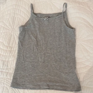 Grå linnetopp med rosett från H&M - Supersöt grå linnetopp från H&M med smala axelband och en liten rosett framtill. Toppen är enkel och stilren, perfekt att matcha med jeans eller kjol. Tillverkad i mjuk bomull för en skön känsla hela dagen.