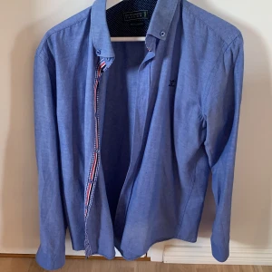 Blå skjorta Sir Raymond Tailor L - Blå långärmad skjorta från Sir Raymond Tailor i regular fit. Skjortan har snygga detaljer med rutigt och randigt tyg på insidan av knappslån samt prickigt tyg i kragen. Tillverkad i 85% bomull och 15% polyester. Perfekt för dig som gillar klassisk stil med en twist.