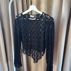 Svart spetsblus  - Säljer en svart långärmad blus från Rita Ora i transparent spets. Blusen har rund halsringning, vågig nederkant och är figurnära. Perfekt för dig som vill ha en edgy och snygg look med mycket detaljer.