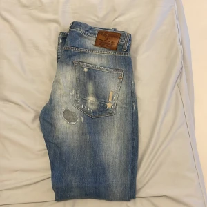 Blå jeans från Reign Italia med patch - Snygga blå jeans från Reign Italia med slitningar, patchdetaljer och broderad stjärna på bakfickan. Jeansen har en klassisk femficksdesign och coolt tvättad look. Perfekta för dig som gillar streetstyle och unika detaljer.