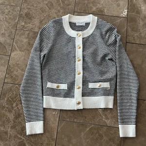 Randig kofta från Holly & Whyte - Snygg randig kofta från Holly & Whyte med svarta och vita ränder, guldiga knappar och ribbade muddar. Koftan har två dekorativa fickor framtill och är långärmad med klassisk rund hals. Perfekt för att lyfta din outfit med en preppy touch.