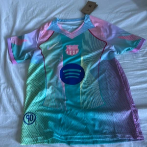 FC Barcelona x Nike färgglad fotbollströja - Säljer en limited edition FC Barcelona x Nike fotbollströja i Dri-FIT material. Tröjan har korta ärmar och ett unikt mönster i turkos, rosa, blått och lila med tunna ränder och prickar. Barcelona-logga och Nike Swoosh på bröstet samt Spotify-logga. Perfekt för dig som vill sticka ut på planen.