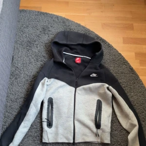 Nike svart och grå hoodie - Nike hoodie i svart och ljusgrått med dragkedja framtill och två fickor med svarta dragkedjor. Jackan har huva och sportig design med logga på bröstet. Perfekt för en chill och aktiv stil.