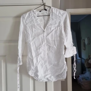 Vit linnehenley från Zara Basic - Vit henley-skjorta från Zara Basic i luftigt linne. Skjortan har v-ringning, knappar framtill, bröstficka och trekvartsärmar med snörning vid ärmslut. Perfekt för en fräsch och avslappnad look.