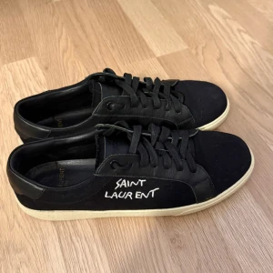 Svarta Saint Laurent sneakers - Snygga svarta sneakers från Saint Laurent med vit logga på sidan och klassisk vit sula. Skorna har snörning, rund tå och är tillverkade i en mix av canvas och skinn. Perfekta för dig som gillar stilrena och exklusiva sneakers med streetkänsla.
